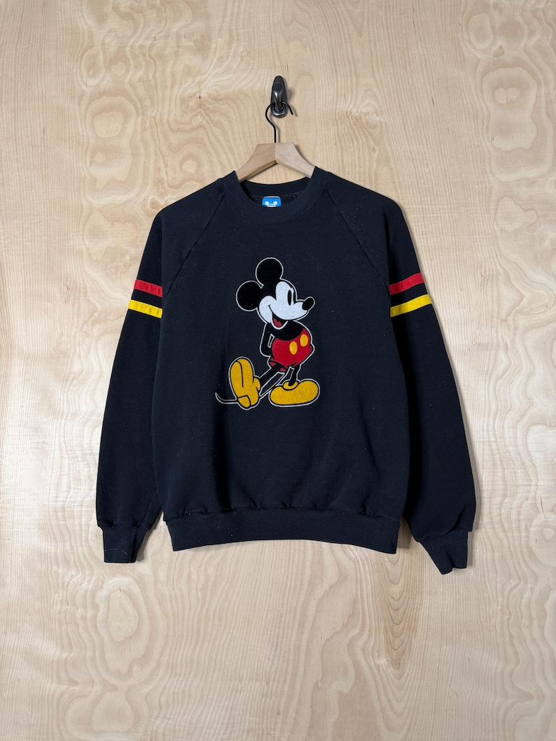 May include: Black sweatshirt featuring a classic Mickey Mouse graphic. The long sleeves have yellow and red stripes. The cartoon character is depicted in his signature outfit with yellow shoes, red shorts, and white gloves.