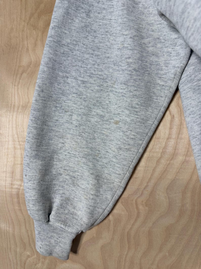 K&ouml;nnte beinhalten: Nahaufnahme eines hellgrauen Sweatshirt-&Auml;rmels. Der &Auml;rmel besteht aus einem weichen, strukturierten Stoff und hat ein geripptes B&uuml;ndchen. Auf dem &Auml;rmel befindet sich ein kleiner Fleck. Das Sweatshirt scheint Vintage oder gebraucht zu sein.