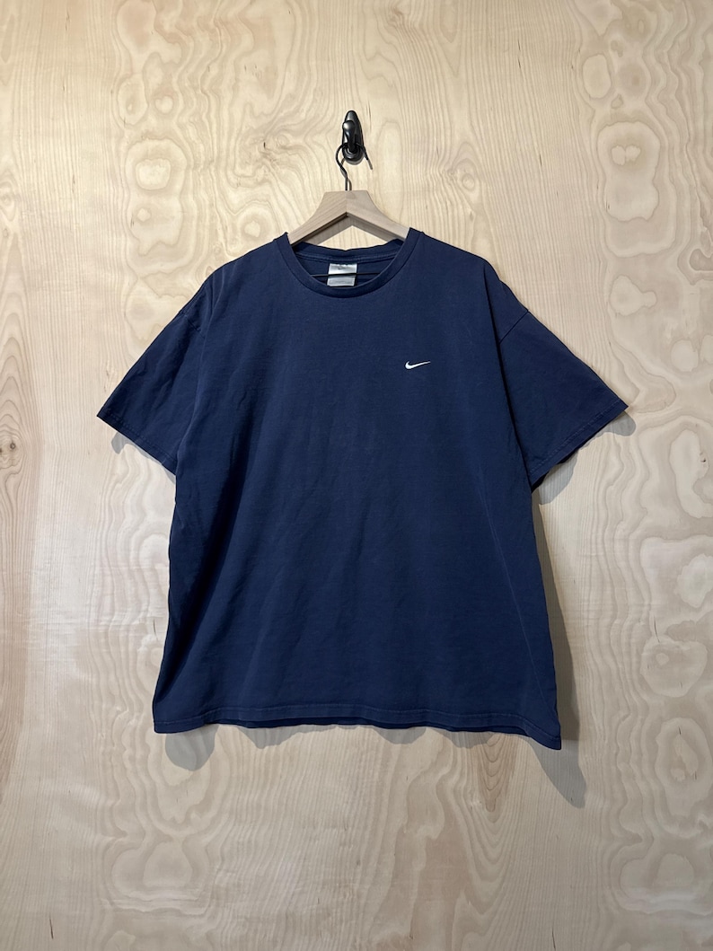 May include: Navy blue t-shirt with a small white Nike logo on the chest. The short-sleeved shirt is hanging on a wooden hanger against a light wood-grain background. The shirt is a solid colour and appears to be made of cotton.