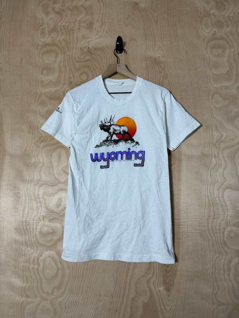 May include: White t-shirt featuring a graphic of a moose silhouette against a setting sun, with the word "Wyoming" in blue and orange. The shirt is short-sleeved and hangs on a wooden hanger.