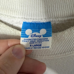 May include: Close-up of a white sweatshirt tag with the Disney logo and text. The blue tag reads "CHARACTER FASHIONS, WALT DISNEY PRODUCTIONS, MADE IN U.S.A., X-LARGE". The sweatshirt is a neutral color.