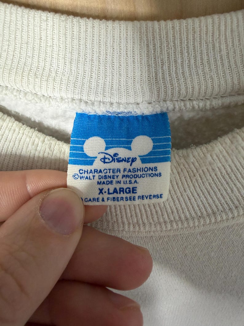 May include: Close-up of a white sweatshirt tag with the Disney logo and text. The blue tag reads "CHARACTER FASHIONS, WALT DISNEY PRODUCTIONS, MADE IN U.S.A., X-LARGE". The sweatshirt is a neutral color.