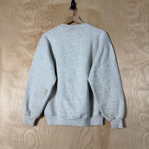 K&ouml;nnte beinhalten: Ein melierter grauer Sweatshirt mit Rundhalsausschnitt und langen &Auml;rmeln, an einem Holzb&uuml;gel aufgeh&auml;ngt. Die R&uuml;ckseite des Kleidungsst&uuml;cks ist sichtbar und zeigt ein schlichtes, l&auml;ssiges Design. Auf dem linken &Auml;rmel befinden sich einige Flecken.