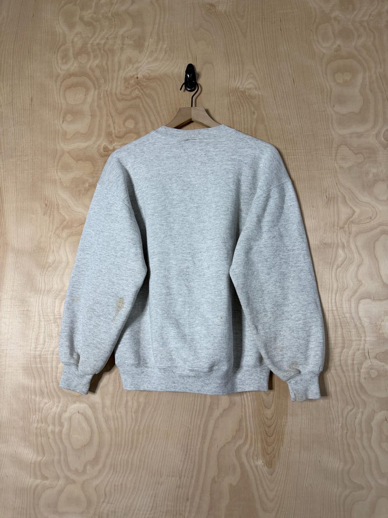 K&ouml;nnte beinhalten: Ein melierter grauer Sweatshirt mit Rundhalsausschnitt und langen &Auml;rmeln, an einem Holzb&uuml;gel aufgeh&auml;ngt. Die R&uuml;ckseite des Kleidungsst&uuml;cks ist sichtbar und zeigt ein schlichtes, l&auml;ssiges Design. Auf dem linken &Auml;rmel befinden sich einige Flecken.