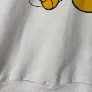 May include: White sweatshirt featuring a partial graphic of Mickey Mouse's yellow gloved hand and foot. The graphic is outlined in black. The sweatshirt has a ribbed hem and cuffs. The fabric appears to be a soft, slightly worn material.