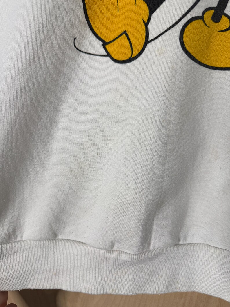 May include: White sweatshirt featuring a partial graphic of Mickey Mouse's yellow gloved hand and foot. The graphic is outlined in black. The sweatshirt has a ribbed hem and cuffs. The fabric appears to be a soft, slightly worn material.