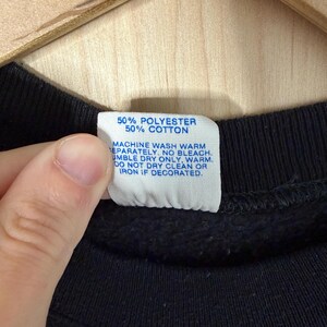 May include: Close-up of a black garment with a white tag. The tag reads "50% POLYESTER 50% COTTON" and washing instructions. The garment is held by a hand, and the background is a light-colored wooden surface.