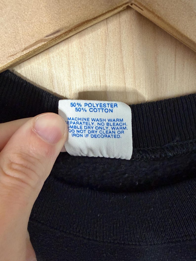 May include: Close-up of a black garment with a white tag. The tag reads "50% POLYESTER 50% COTTON" and washing instructions. The garment is held by a hand, and the background is a light-colored wooden surface.