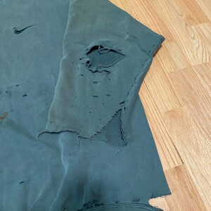 May include: A dark green sweatshirt with a Nike logo on the left chest. The sweatshirt has several holes and tears throughout, giving it a distressed look.