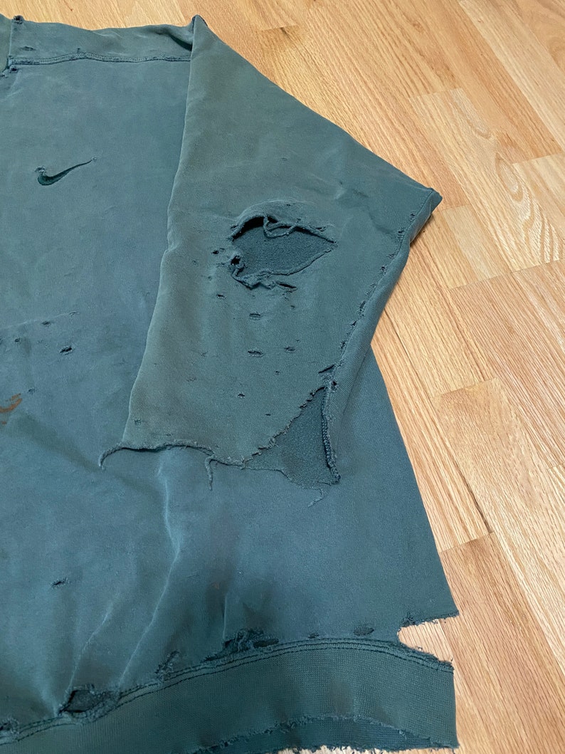 May include: A dark green sweatshirt with a Nike logo on the left chest. The sweatshirt has several holes and tears throughout, giving it a distressed look.