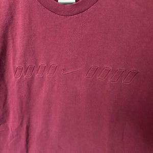 K&ouml;nnte beinhalten: Burgunderfarbenes T-Shirt mit Rundhalsausschnitt und tonalen, gepr&auml;gten Nike-Logo auf der Brust. Das Shirt hat eine klassische Passform und ein Etikett am Nacken.