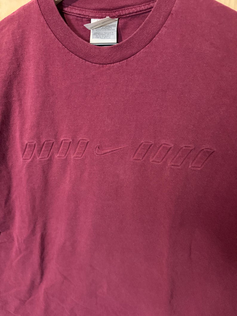 K&ouml;nnte beinhalten: Burgunderfarbenes T-Shirt mit Rundhalsausschnitt und tonalen, gepr&auml;gten Nike-Logo auf der Brust. Das Shirt hat eine klassische Passform und ein Etikett am Nacken.