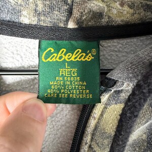 May include: A camouflage print hooded sweatshirt with a green fabric tag featuring the Cabela's logo, size L REG, RN 56835, Made in China, 60% cotton, 40% polyester, and the instruction to see reverse for care details.