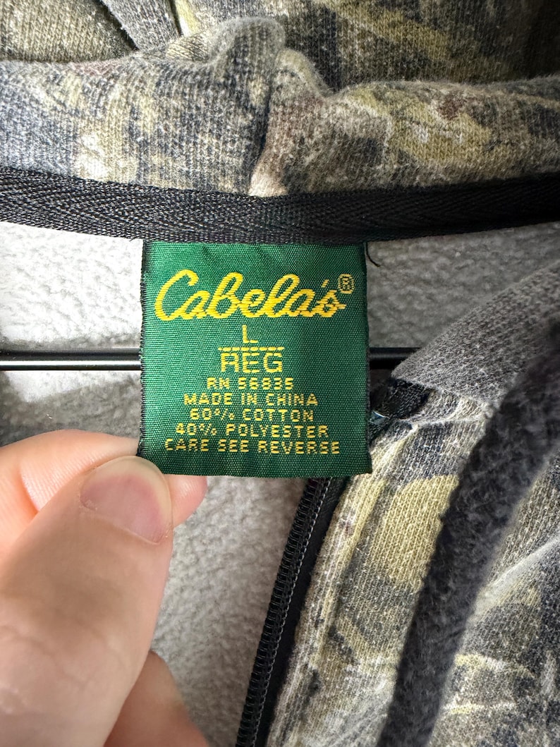 May include: A camouflage print hooded sweatshirt with a green fabric tag featuring the Cabela's logo, size L REG, RN 56835, Made in China, 60% cotton, 40% polyester, and the instruction to see reverse for care details.
