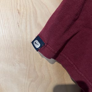 K&ouml;nnte beinhalten: Nahaufnahme eines weinroten T-Shirt-&Auml;rmels. Der &Auml;rmel hat ein kleines, rechteckiges Etikett mit dem wei&szlig;en Nike Swoosh-Logo auf marineblauem Hintergrund. Der Stoff scheint ein weiches Strickmaterial zu sein.