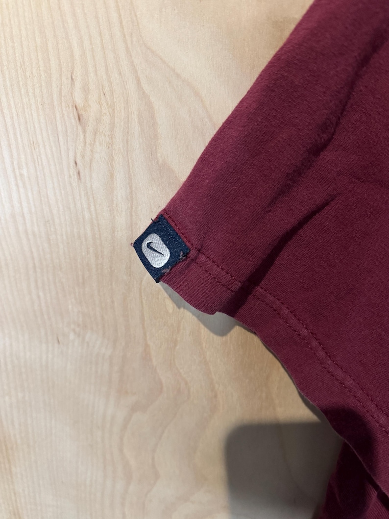 K&ouml;nnte beinhalten: Nahaufnahme eines weinroten T-Shirt-&Auml;rmels. Der &Auml;rmel hat ein kleines, rechteckiges Etikett mit dem wei&szlig;en Nike Swoosh-Logo auf marineblauem Hintergrund. Der Stoff scheint ein weiches Strickmaterial zu sein.