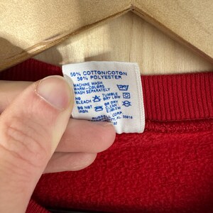 May include: A close-up of a red sweatshirt's label, detailing fabric composition and care instructions. The label states "50% Cotton/Coton, 50% Polyester" and features washing symbols. The sweatshirt is suspended on a wooden hanger.