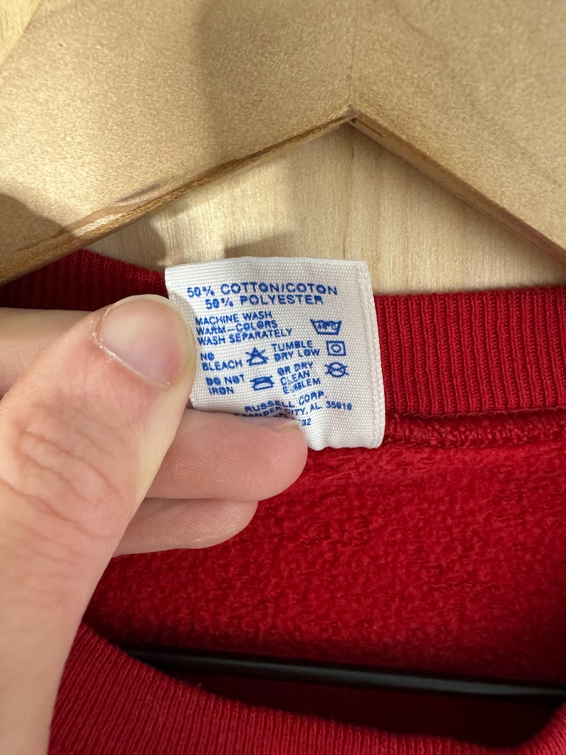 May include: A close-up of a red sweatshirt's label, detailing fabric composition and care instructions. The label states "50% Cotton/Coton, 50% Polyester" and features washing symbols. The sweatshirt is suspended on a wooden hanger.