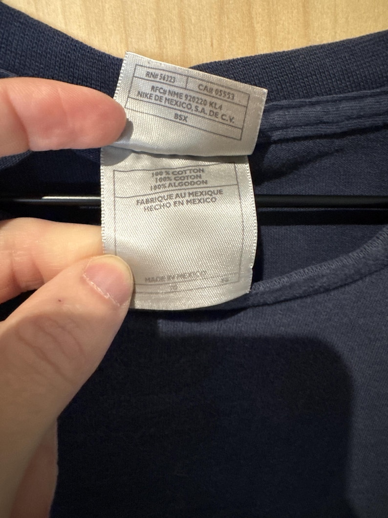 May include: Close-up of a navy blue t-shirt with a tag attached. The tag includes text such as "100% COTTON" and "MADE IN MEXICO". The tag also includes the text "NIKE DE MEXICO, S.A. DE C.V."