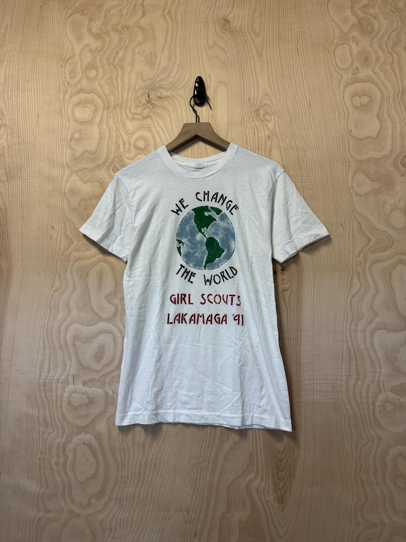 Peut inclure: T-shirt blanc avec un motif de globe terrestre et l'inscription "WE CHANGE THE WORLD". En dessous, on peut lire "GIRL SCOUTS LAKAMAGA '91" en rouge. Le t-shirt est suspendu &agrave; un cintre en bois.