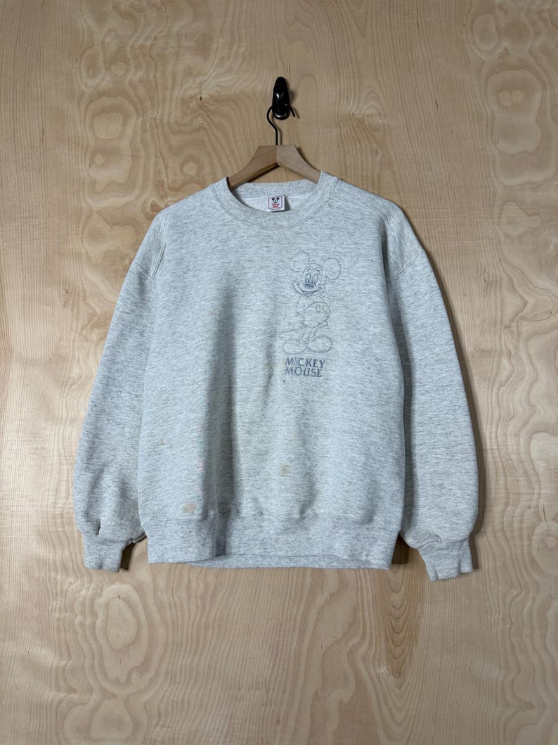 K&ouml;nnte beinhalten: Ein hellgraues Sweatshirt mit einer Mickey Mouse-Grafik. Die Grafik ist eine Strichzeichnung von Mickey Mouse mit den Worten "Mickey Mouse" darunter. Das Sweatshirt hat einen Rundhalsausschnitt und lange &Auml;rmel.