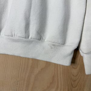 May include: Close-up of a white sweatshirt. The ribbed hem and cuffs are visible, along with the soft, textured fabric. The sweatshirt appears to be slightly worn, with subtle discoloration. The background is a light-colored wooden surface.