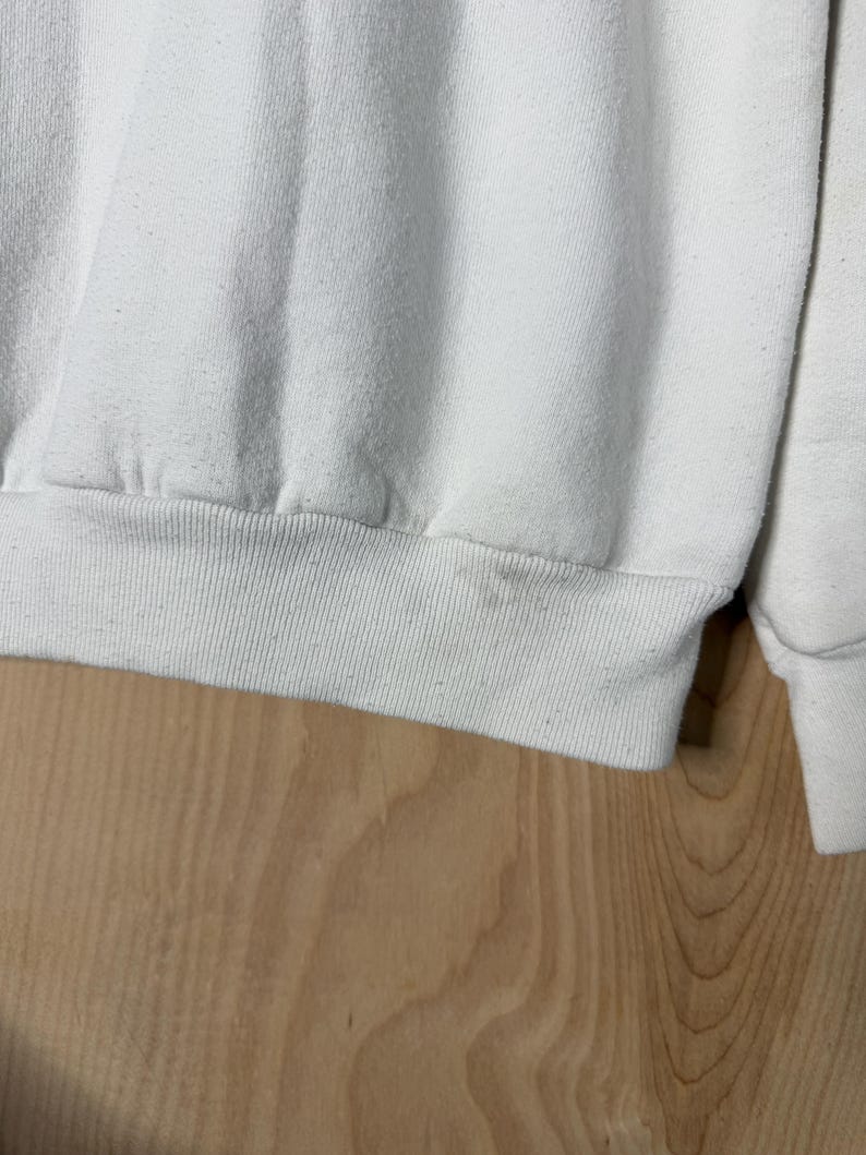 May include: Close-up of a white sweatshirt. The ribbed hem and cuffs are visible, along with the soft, textured fabric. The sweatshirt appears to be slightly worn, with subtle discoloration. The background is a light-colored wooden surface.