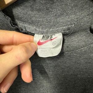 May include: A gray Nike t-shirt with a white tag that says "Nike" in black lettering and a white swoosh logo. The tag also says "XL" in black lettering.