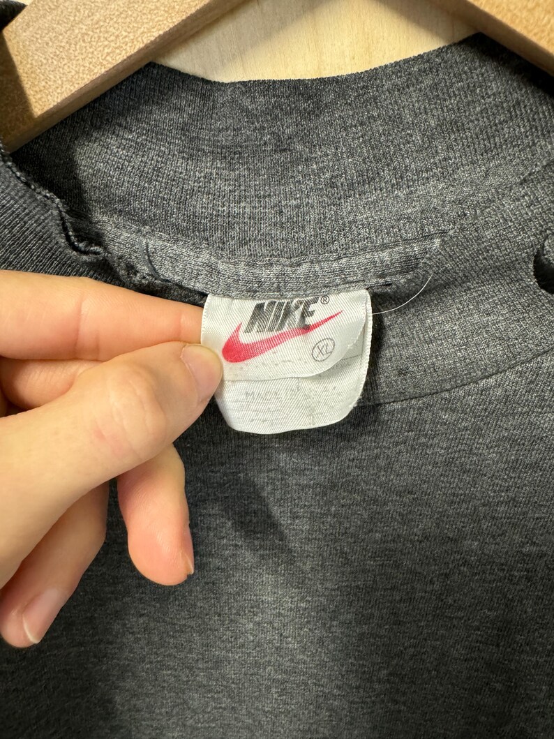 May include: A gray Nike t-shirt with a white tag that says "Nike" in black lettering and a white swoosh logo. The tag also says "XL" in black lettering.