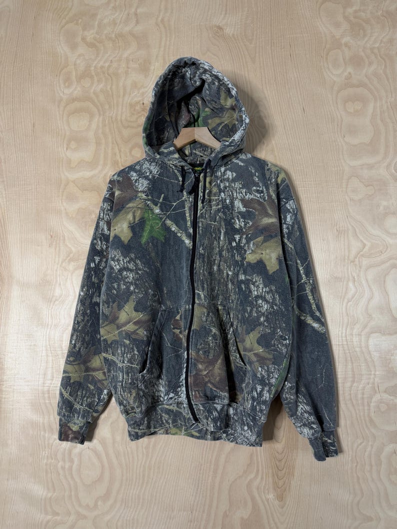 May include: A long-sleeve zip-up hoodie featuring a realistic forest camouflage pattern in shades of dark grey, black, and muted green. The garment includes a hood with black drawstrings, a full-length front zip, and two front hand pockets.