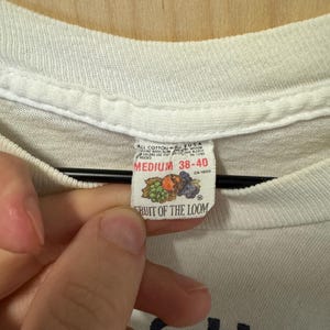 Peut inclure: Gros plan sur une &eacute;tiquette de t-shirt blanche. L'&eacute;tiquette indique "FRUIT OF THE LOOM" avec un graphique de fruits. L'&eacute;tiquette indique &eacute;galement la taille "MEDIUM 38-40". Le t-shirt est en coton.