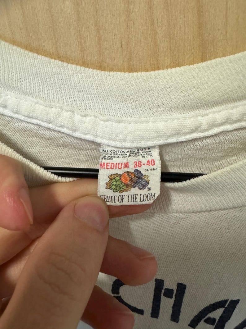Peut inclure: Gros plan sur une &eacute;tiquette de t-shirt blanche. L'&eacute;tiquette indique "FRUIT OF THE LOOM" avec un graphique de fruits. L'&eacute;tiquette indique &eacute;galement la taille "MEDIUM 38-40". Le t-shirt est en coton.