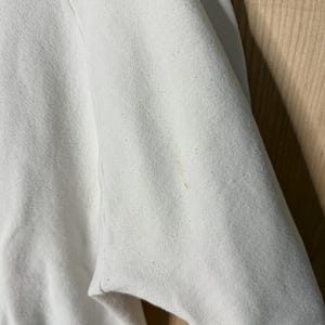 May include: Close-up of a white sweatshirt sleeve. The fabric is slightly textured with small flecks. A small, light brown stain is visible on the sleeve. The sleeve is partially folded, revealing the inside of the garment.