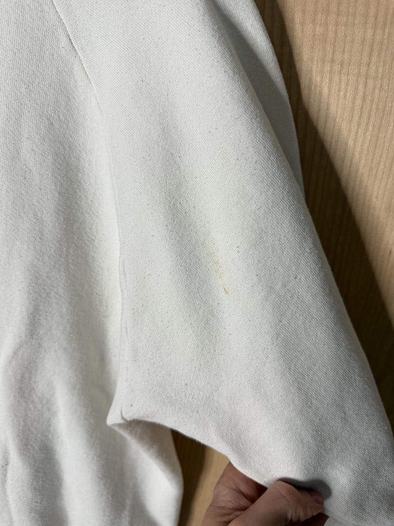 May include: Close-up of a white sweatshirt sleeve. The fabric is slightly textured with small flecks. A small, light brown stain is visible on the sleeve. The sleeve is partially folded, revealing the inside of the garment.