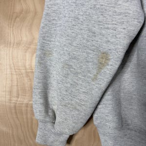 K&ouml;nnte beinhalten: Nahaufnahme eines hellgrauen Sweatshirt-&Auml;rmels mit geripptem B&uuml;ndchen. Der &Auml;rmel hat mehrere braune Flecken. Das Sweatshirt besteht aus einem weichen, strukturierten Stoff. Der Hintergrund ist eine helle Holzmaserung.