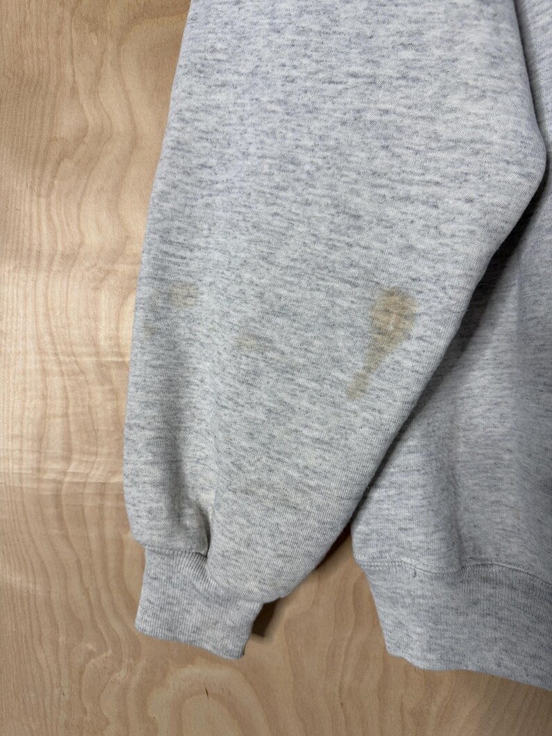 K&ouml;nnte beinhalten: Nahaufnahme eines hellgrauen Sweatshirt-&Auml;rmels mit geripptem B&uuml;ndchen. Der &Auml;rmel hat mehrere braune Flecken. Das Sweatshirt besteht aus einem weichen, strukturierten Stoff. Der Hintergrund ist eine helle Holzmaserung.