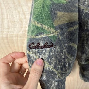 May include: A long-sleeved sweatshirt featuring a realistic forest camouflage pattern in shades of green, brown, and grey. The word Cabela's is embroidered in dark brown thread on the lower portion of the sleeve near the ribbed cuff.