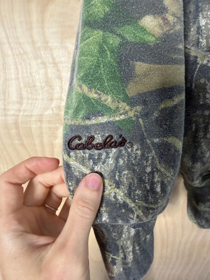 May include: A long-sleeved sweatshirt featuring a realistic forest camouflage pattern in shades of green, brown, and grey. The word Cabela's is embroidered in dark brown thread on the lower portion of the sleeve near the ribbed cuff.