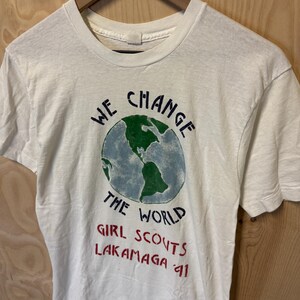 Peut inclure: T-shirt blanc avec un motif de globe terrestre et l'inscription "WE CHANGE THE WORLD" en bleu et noir. En dessous, le texte "GIRL SCOUTS LAKAMAGA 91" en rouge. Le t-shirt est suspendu &agrave; un cintre en bois.