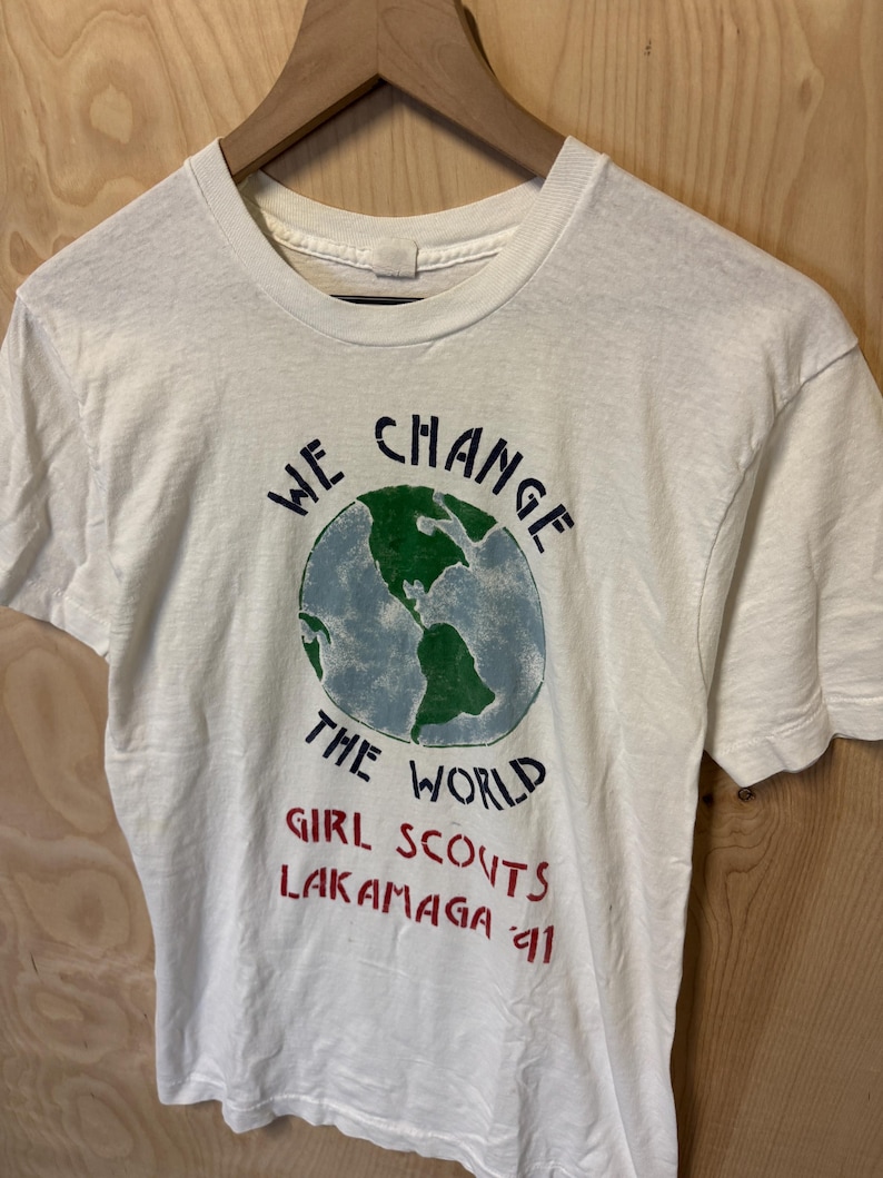 Peut inclure: T-shirt blanc avec un motif de globe terrestre et l'inscription "WE CHANGE THE WORLD" en bleu et noir. En dessous, le texte "GIRL SCOUTS LAKAMAGA 91" en rouge. Le t-shirt est suspendu &agrave; un cintre en bois.
