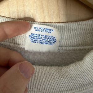 May include: Close-up of a gray sweatshirt collar with a tag. The tag reads "50% POLYESTER 50% COTTON" and washing instructions. The sweatshirt is made of a soft, textured fabric.