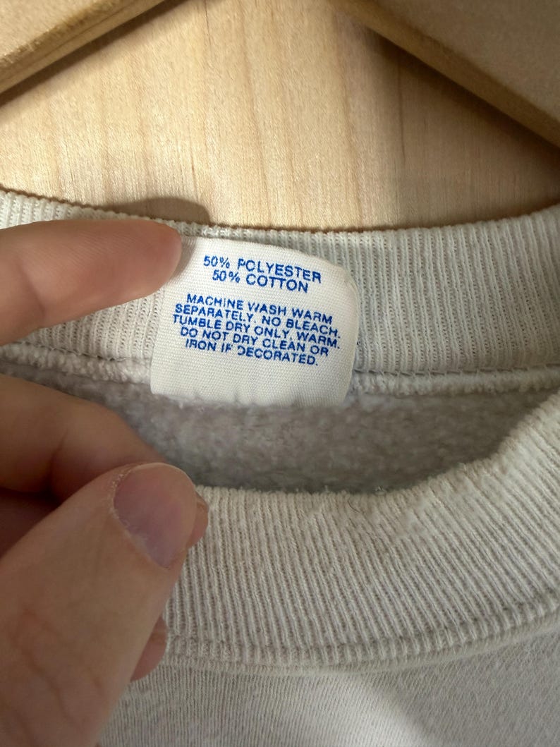 May include: Close-up of a gray sweatshirt collar with a tag. The tag reads "50% POLYESTER 50% COTTON" and washing instructions. The sweatshirt is made of a soft, textured fabric.
