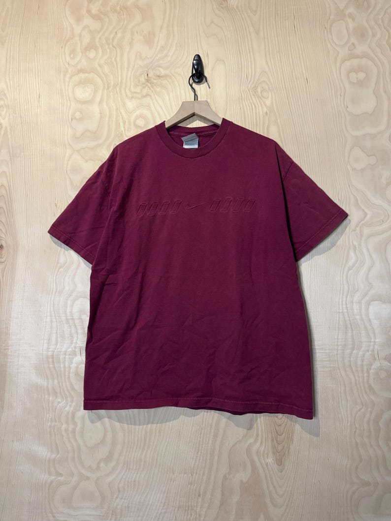 K&ouml;nnte beinhalten: Ein weinrotes T-Shirt mit kurzen &Auml;rmeln, das an einem Holzb&uuml;gel h&auml;ngt. Das Shirt hat den gepr&auml;gten Text "good - good" auf der Brust. Das Shirt ist aus einem weichen Material.