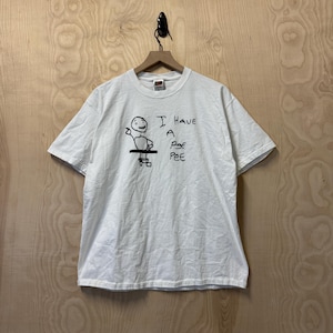 Könnte beinhalten: Weißes T-Shirt mit einer schwarzen, handgezeichneten Grafik einer Strichmännchenfigur und dem Text "I Have A Pee Pee". Das T-Shirt hängt vor einem Holzgrund.