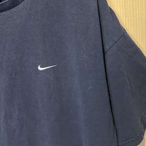 May include: Navy blue t-shirt featuring a white Nike swoosh logo on the chest. The top has short sleeves and a crew neck. The fabric appears slightly faded, giving it a vintage appearance.