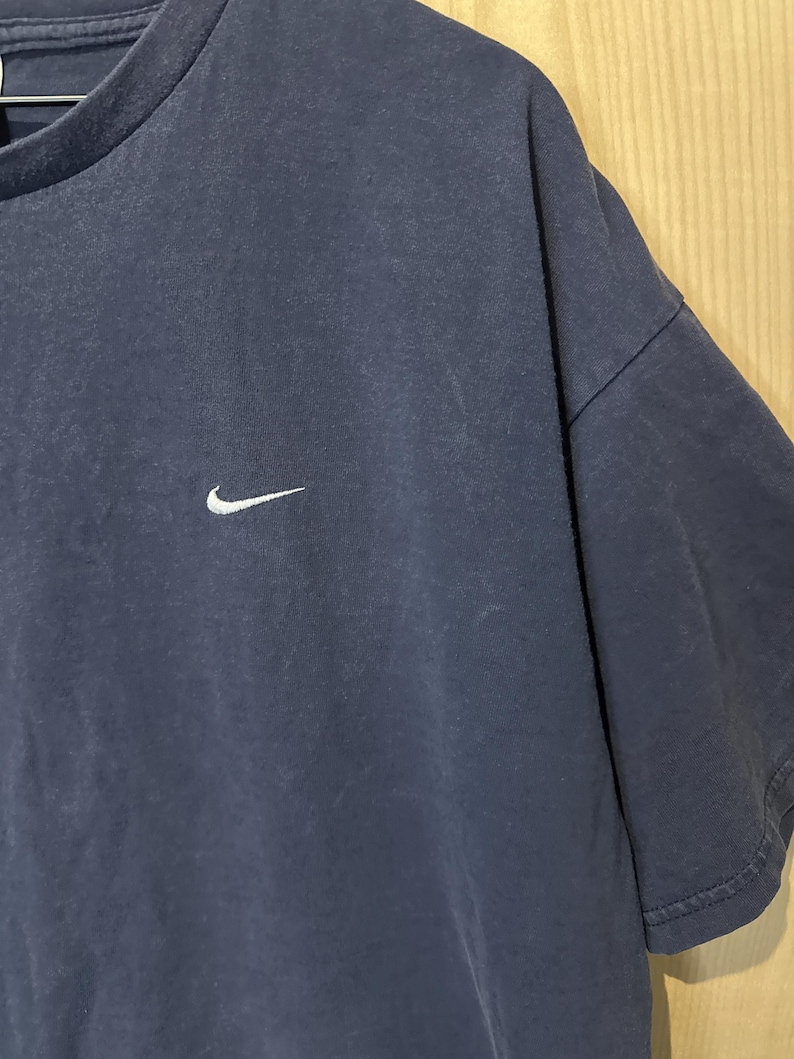 May include: Navy blue t-shirt featuring a white Nike swoosh logo on the chest. The top has short sleeves and a crew neck. The fabric appears slightly faded, giving it a vintage appearance.
