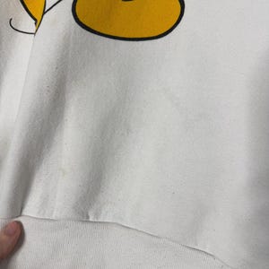 May include: White sweatshirt featuring a partial graphic of Mickey Mouse's yellow shoe and black leg. The sweatshirt has a ribbed hem and appears to be vintage. The graphic is outlined in black.