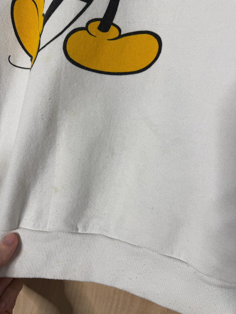 May include: White sweatshirt featuring a partial graphic of Mickey Mouse's yellow shoe and black leg. The sweatshirt has a ribbed hem and appears to be vintage. The graphic is outlined in black.