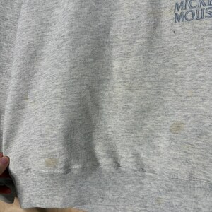 K&ouml;nnte beinhalten: Ein hellgraues Sweatshirt mit dem blauen Aufdruck "Mickey Mouse". Das Sweatshirt hat einen gerippten Saum und B&uuml;ndchen. Auf der Vorderseite des Kleidungsst&uuml;cks sind mehrere Flecken zu sehen.