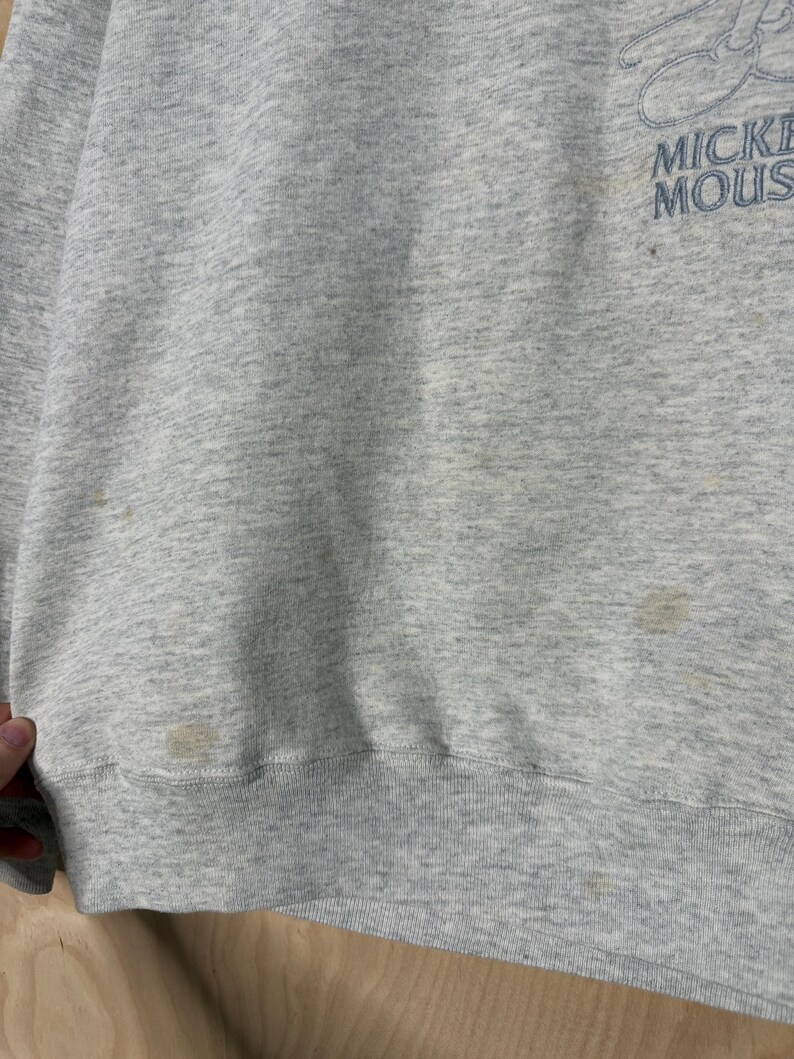 K&ouml;nnte beinhalten: Ein hellgraues Sweatshirt mit dem blauen Aufdruck "Mickey Mouse". Das Sweatshirt hat einen gerippten Saum und B&uuml;ndchen. Auf der Vorderseite des Kleidungsst&uuml;cks sind mehrere Flecken zu sehen.
